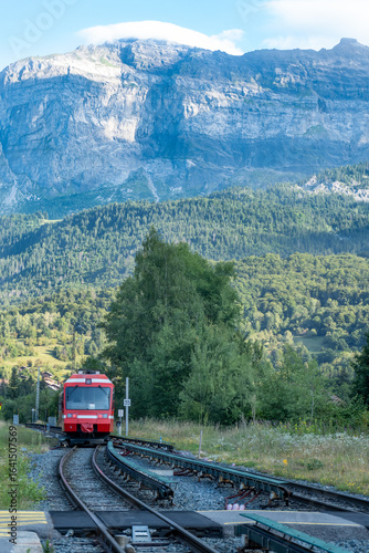 A red train departing Servoz station in the French Alps with green hills and rocky mountains behind.