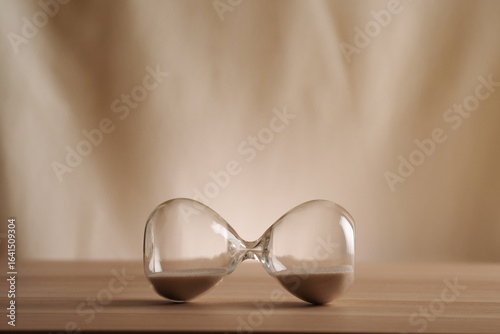 A simple hourglass on it's side on a wooden table with a beige backdrop, symbolizing time flow and minimalistic living. Represents timeless balance in a creative, uncluttered presentation concepts