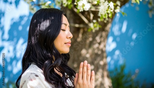 A peaceful and relaxed Asian woman meditating with her hands in prayer, breathing deeply and enjoying the fresh air in a tranquil garden.