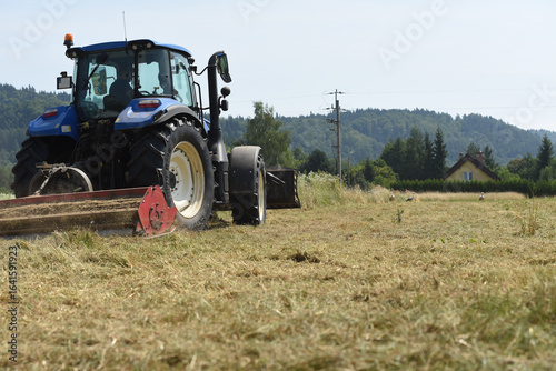 Agricultural tractor mowing grass in a countryside field, with storks visible in the background and a house surrounded by trees