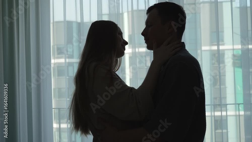 Silhouette of loving couple looking and standing facing each other against background of window. Intimate moment and tender embrace of man and woman in house.