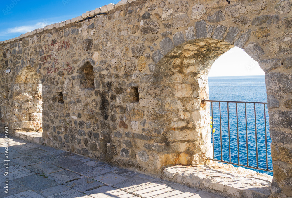 Naklejka premium Stone wall with arched openings in Ulcinj’s Old Town, Montenegro, overlooking the Adriatic Sea. Sunlight illuminates the historic masonry, revealing a scenic coastal view through the arches.