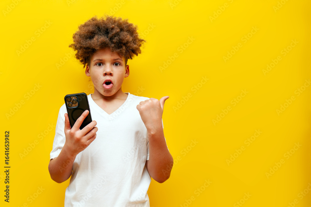 shocked african american boy using smartphone and pointing at copy space on yellow isolated background, surprised schoolboy teenager holding mobile phone and pointing at empty space in amazement shocked african american boy using smartphone and pointing at copy space on yellow isolated background, surprised schoolboy teenager holding mobile phone and pointing at empty space in amazement