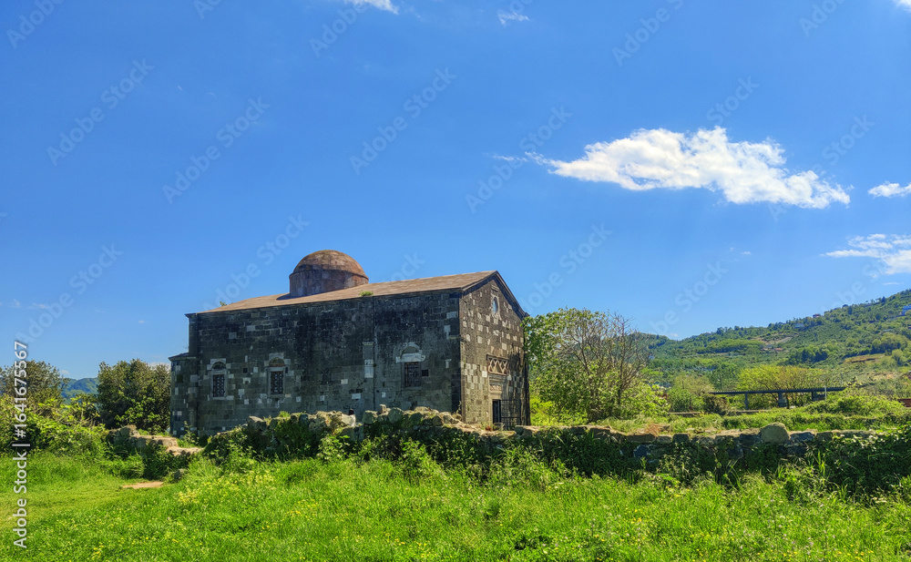 Obraz premium Historic stone church with domed roof. Stone building topped with a domed roof sits behind a low stone wall in a lush green field under a bright blue sky. perfect for travel and architecture.