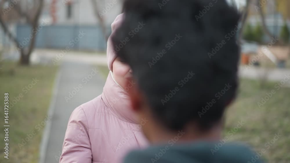 Smiling woman in pink winter jacket and matching beanie with long braid walking outdoors in park on paved path towards person in foreground during early spring with grass and bare trees in background Smiling woman in pink winter jacket and matching beanie with long braid walking outdoors in park on paved path towards person in foreground during early spring with grass and bare trees in background