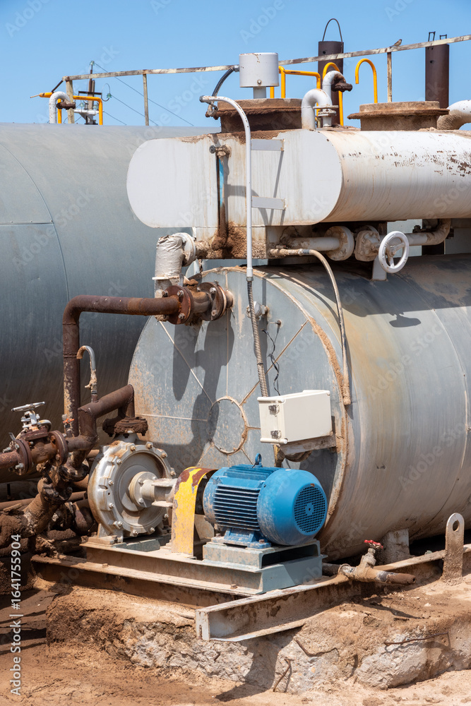 Naklejka premium Industrial oil pump with electric motor connected to weathered metal tank, realistic style, on sandy ground, concept of heavy machinery operation