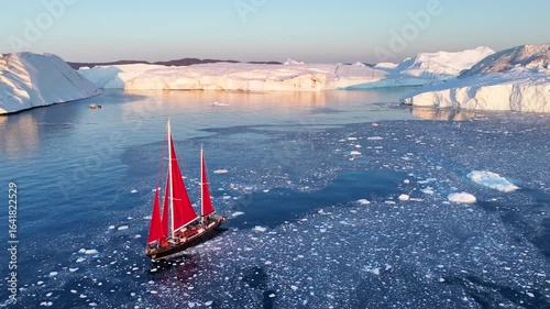 Little red sailboat cruising among floating icebergs in Disko Bay glacier during midnight sun season of polar summer. Ilulissat, Greenland. Global warming and melting glaciers.