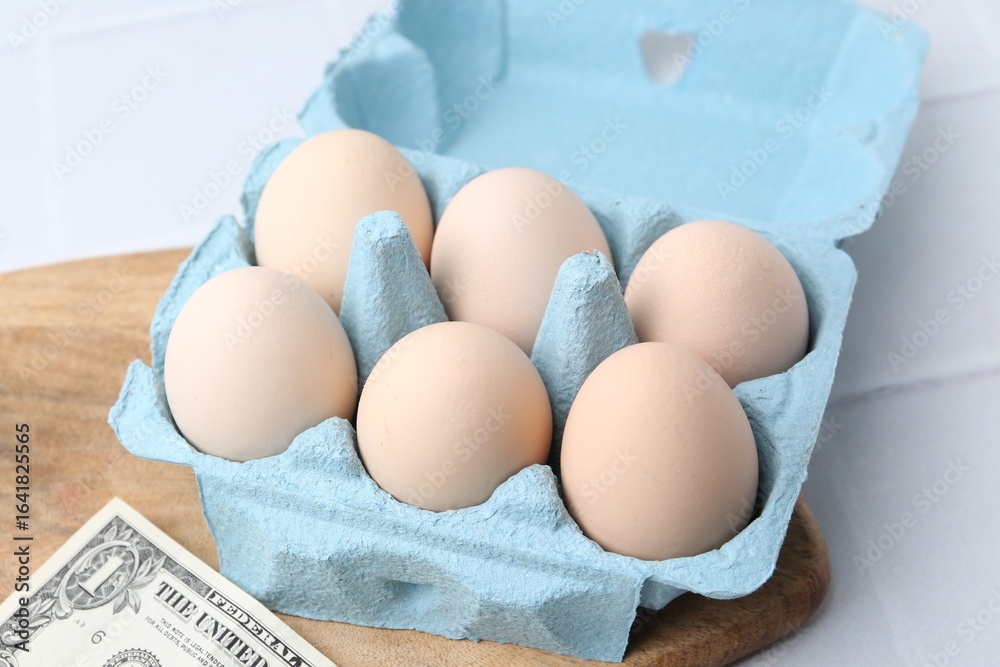 Raw chicken eggs in egg carton and dollar banknote on white tiled table, closeup Raw chicken eggs in egg carton and dollar banknote on white tiled table, closeup