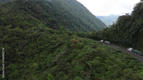 Forward aerial drone shot approaching trucks driving on mountain road surrounded by lush green forest
