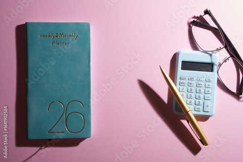 2026 small planner calculator, eyeglasses and pen against pink background.