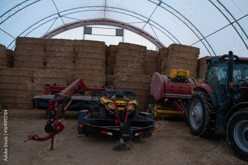 A wide view of various pieces of farming machinery, including a tractor, ATV, and baler, stored next to stacked hay bales inside a hoop barn.