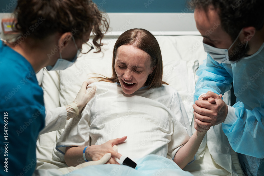 Fototapeta premium Caucasian young adult woman experiencing labor pain in hospital bed, supported by medical professional and Caucasian young adult man holding her hand during childbirth process