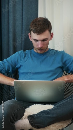 Man is sitting on cozy chair at home and opening laptop computer. Vertical video