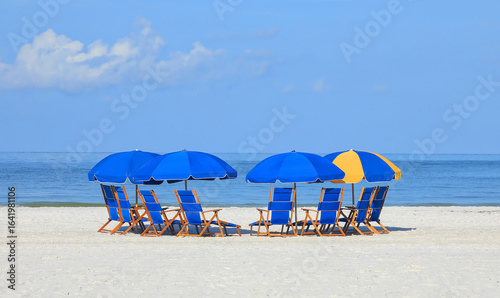Large group of blue folding beach chairs arranged in a semi circle in front of calm blue waters.