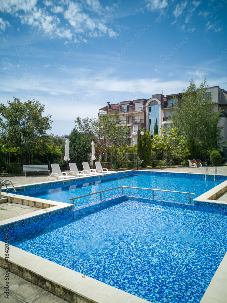 Fototapeta premium Clean outdoor pool with clear blue water, sun loungers, and umbrellas near modern apartment building. Peaceful summer vibe and relaxation area.