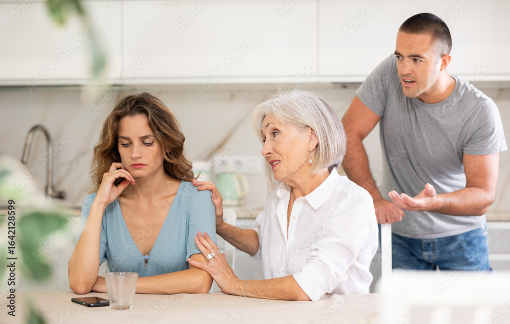 Fototapeta premium Young girl is sitting at table in kitchen quarreling with her mother-in-law and husband