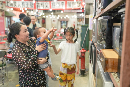 Photography In an old-town-style mall, a Chinese family enjoys the day; a one-year-old in a stroller and kids under ten chat as adults in their thirties gather to watch old televisions