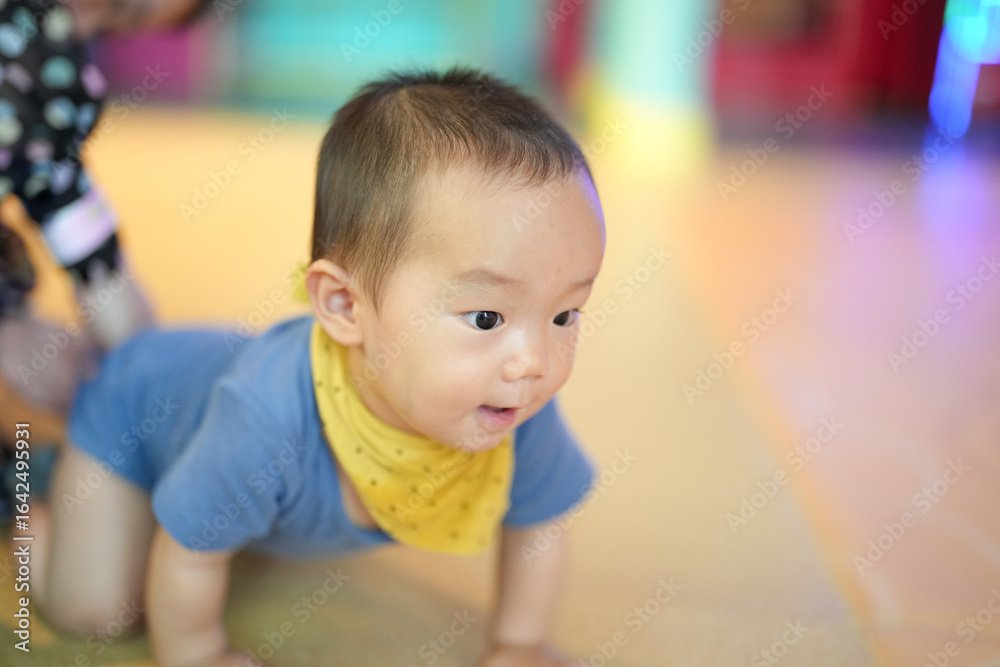 Fototapeta premium In an indoor play area, parents in their thirties watch a one-year-old baby crawl on a soft mat with warm smiles, toys nearby and gentle light in the room. Hongkou District, Shanghai, China. Spring.