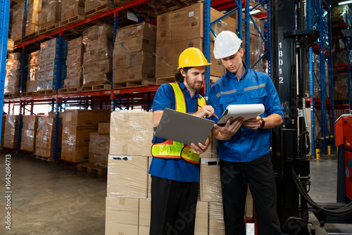 Warehouse supervisor and worker discussing inventory checklist using laptop and clipboard in large distribution center. Efficient teamwork in industrial supply chain logistics.