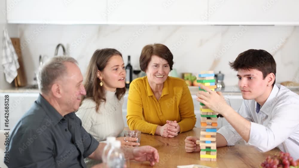 Young guy playing jenga with family in cozy home kitchen, trying to put brick without destroying tower, under interested glances of wife and elderly parents sitting at table with glasses of wine. High