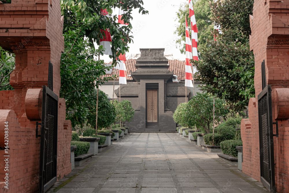 Obraz premium View through a red brick gateway to a classic Javanese house, with a path lined by Indonesian national flags in a green garden setting.