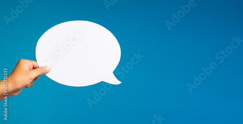 Hand holding blank round speech bubble cutout on blue background for communication and dialogue concept.