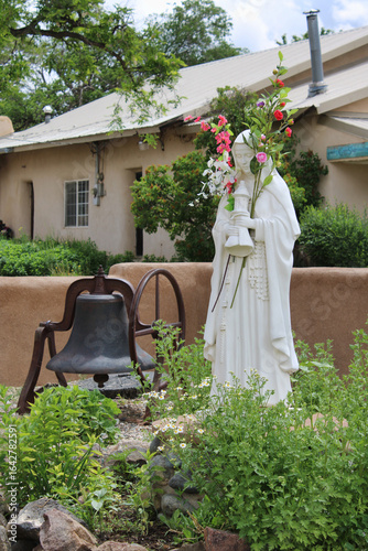 Foto Statue of Virgin Mary Standing in Garden and Holding Flowers Near Large Bell in
