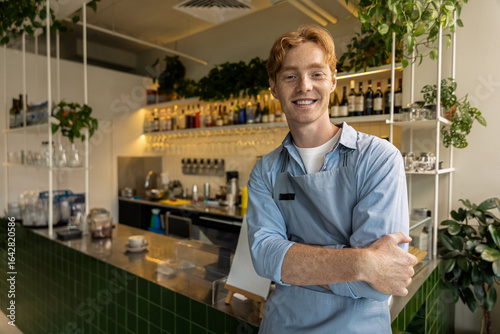 Cheerful young café worker with crossed arms leaning against counter in stylish interior