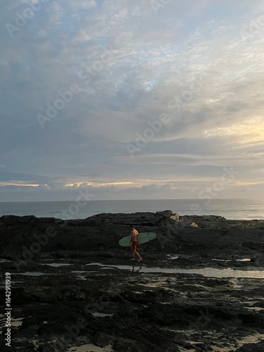 Schilderij op canvas A lone surfer walks across dark coastal rocks at dawn, surfboard in hand, ready for the first waves of the day