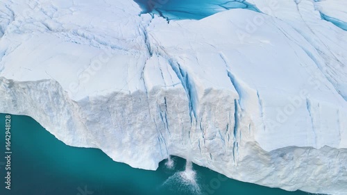 A tall melting iceberg in the Arctic Ocean. A glacier with a lake and waterfall. Clear ice floats in turquoise water. Ecology, melting ice, climate change, global warming. Aerial view. Midnight sun.