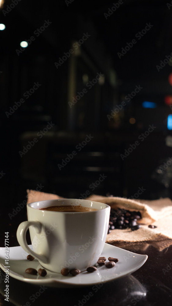 Fototapeta premium A white ceramic cup filled with rich black coffee, placed on a square white saucer with scattered coffee beans, set on a dark reflective surface with a blurred background of coffee beans on burlap.