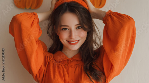 An adult woman wearing a bright orange pumpkin dress with a playful smile and simple makeup.