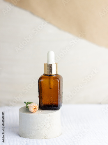 An elegant bottle in noble dark glass with natural serum and the most delicate cream to brighten your skin is decorated with an elegant flower on a plaster catwalk.