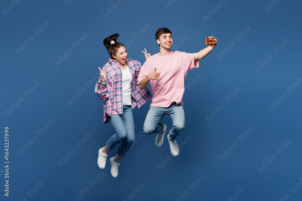 Full body young couple two friends family man woman wear pink casual clothes together jump high doing selfie shot on mobile cell phone show v-sign isolated on plain blue background. Lifestyle concept. Full body young couple two friends family man woman wear pink casual clothes together jump high doing selfie shot on mobile cell phone show v-sign isolated on plain blue background. Lifestyle concept.