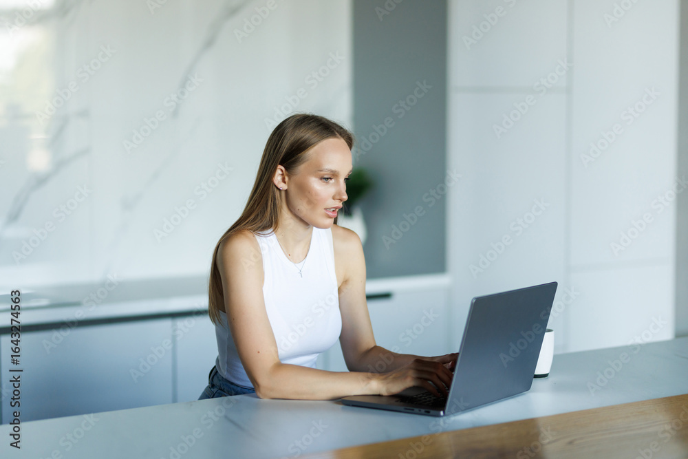 Fototapeta premium Young woman is sitting at the kitchen table with laptop