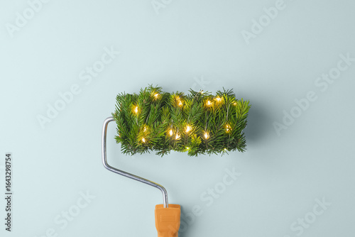 Fototapeta Naklejka Na Ścianę i Meble -  A paint roller wrapped in spruce with warm fairy lights promises a festive home makeover on a soft blue background this winter.