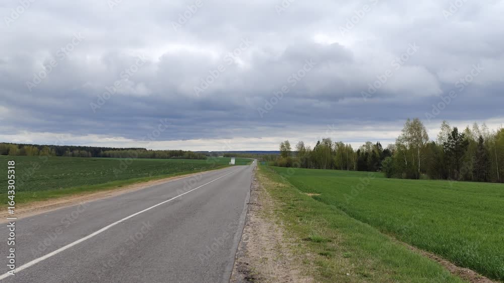 The asphalt road with markings, sandy roadsides and with a bus stop passes among mixed forests and agricultural fields with young green sprouts. Cars are driving Overcast spring weather and cloudy sky The asphalt road with markings, sandy roadsides and with a bus stop passes among mixed forests and agricultural fields with young green sprouts. Cars are driving Overcast spring weather and cloudy sky