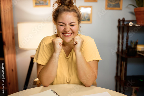 Indoor image of super excited female with hair bun doing winner gesture with clenched fists and closed eyes sitting at table at home, achieving long-term goal passing exam or getting new job