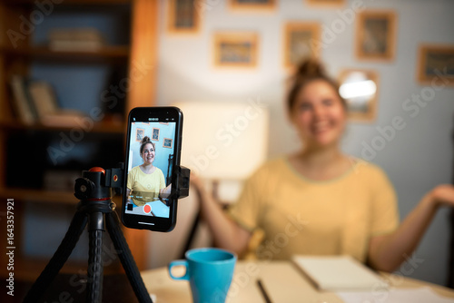 Selective focus of smartphone on tripod with image of Caucasian female English teacher recording video tutorial to get new followers and clients sitting in her cabinet at home gesticulating