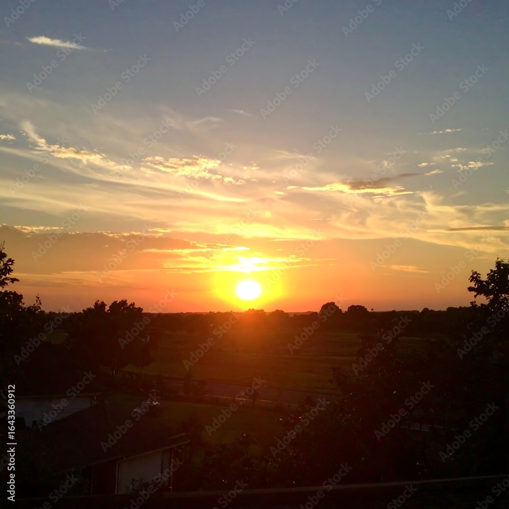 Naklejka premium Sunset over a flat landscape, silhouetting trees and houses