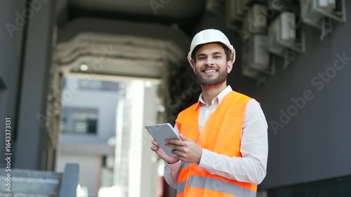Portrait of smiling professional engineer wearing safety helme, vest standing at factory holding using digital tablet in hands. Headshot of confident handsome industry manager posing looking at camera