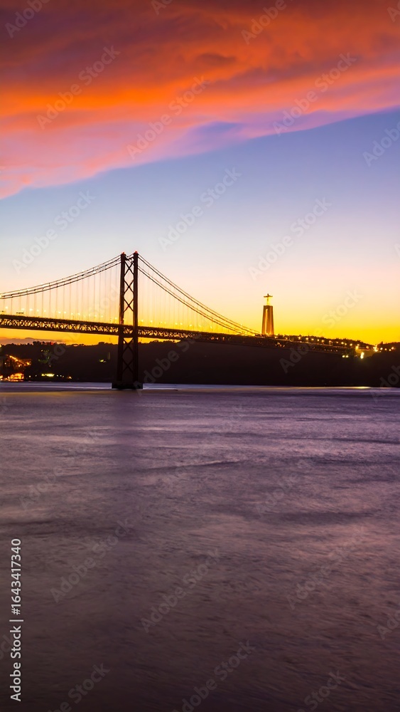 Fototapeta premium Vibrant sunset hues illuminate a suspension bridge and cityscape across a calm river