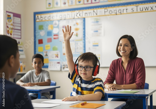 Autistic child boy with red noise cancelling headphones raising hand in inclusive classroom during interactive lesson with diverse students and smiling teacher