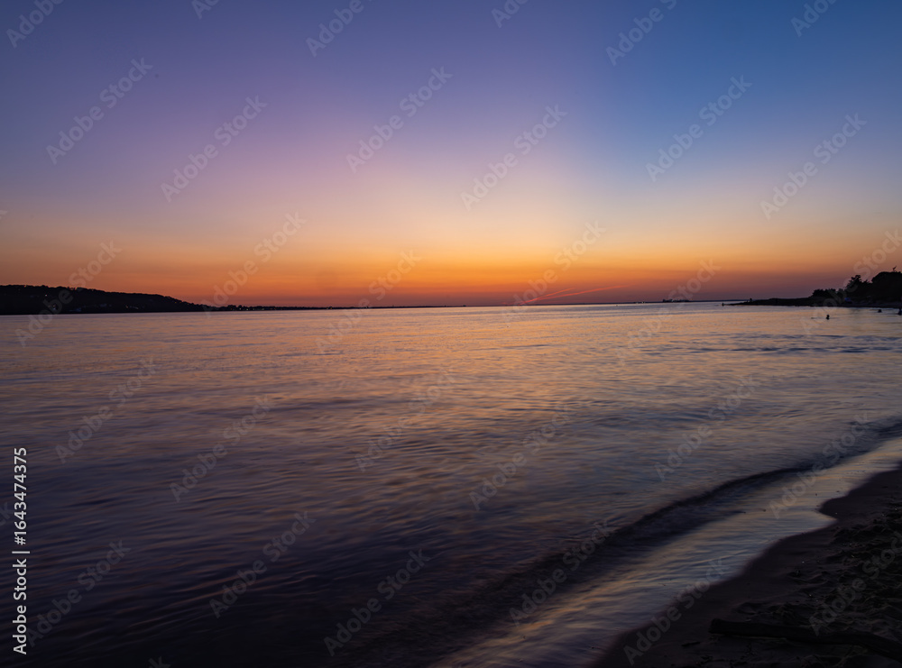 Colorful sunset over calm ocean waters with soft waves lapping the sandy shore, a warm orange and purple sky, and a peaceful horizon creating a tranquil summer coastal scene. Colorful sunset over calm ocean waters with soft waves lapping the sandy shore, a warm orange and purple sky, and a peaceful horizon creating a tranquil summer coastal scene.