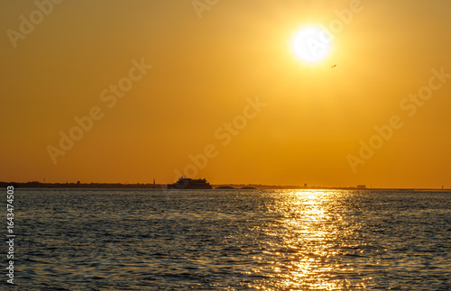 Golden sunset over the sea with shimmering reflections on gentle waves, a distant coastline, and warm orange sky, capturing the serene beauty of a peaceful summer evening by the coast.