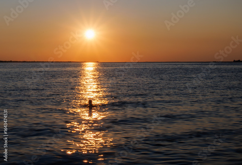 Silhouette of a person swimming in calm sea waters during a golden sunset, with the sun casting a warm reflection across gentle waves under an orange evening sky.