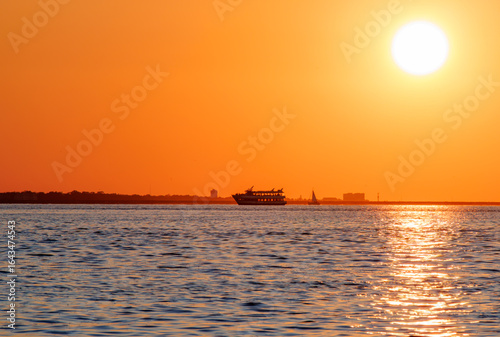 Silhouette of a boat sailing across calm waters during a vivid orange sunset, with the sun casting golden reflections and creating a warm, tranquil maritime evening scene.