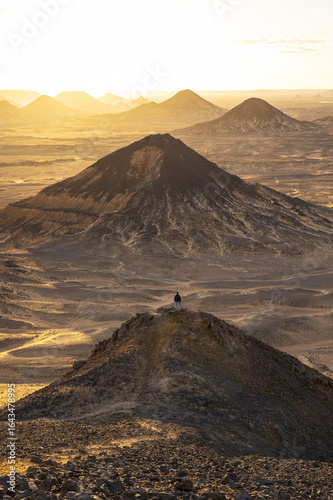 View of the sun-kissed desert landscape unfolds with layered mountains and a lone figure perched atop the foremost peak, Bawati, Giza Governorate, Egypt.