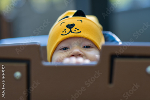 Baby wearing a yellow dog hat peeking out from a play structure in a park during daytime