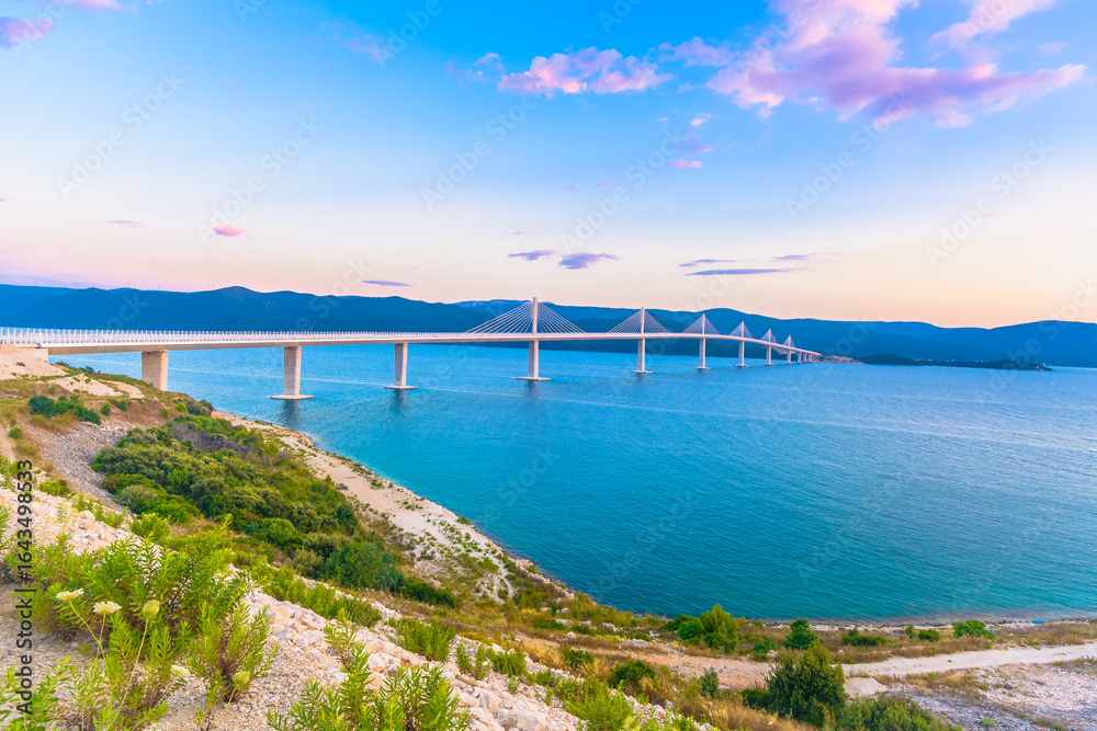 Obraz premium Scenic view of the Peljesac Bridge stretching across the blue Adriatic Sea, with its modern white pillars and surrounding coastal landscape in southern Croatia under clear sunny skies